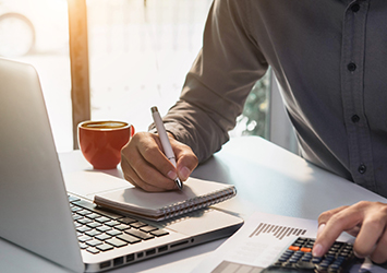 Close up of person at a desk with a laptop, typing on a calculator and writing with a pen and paper